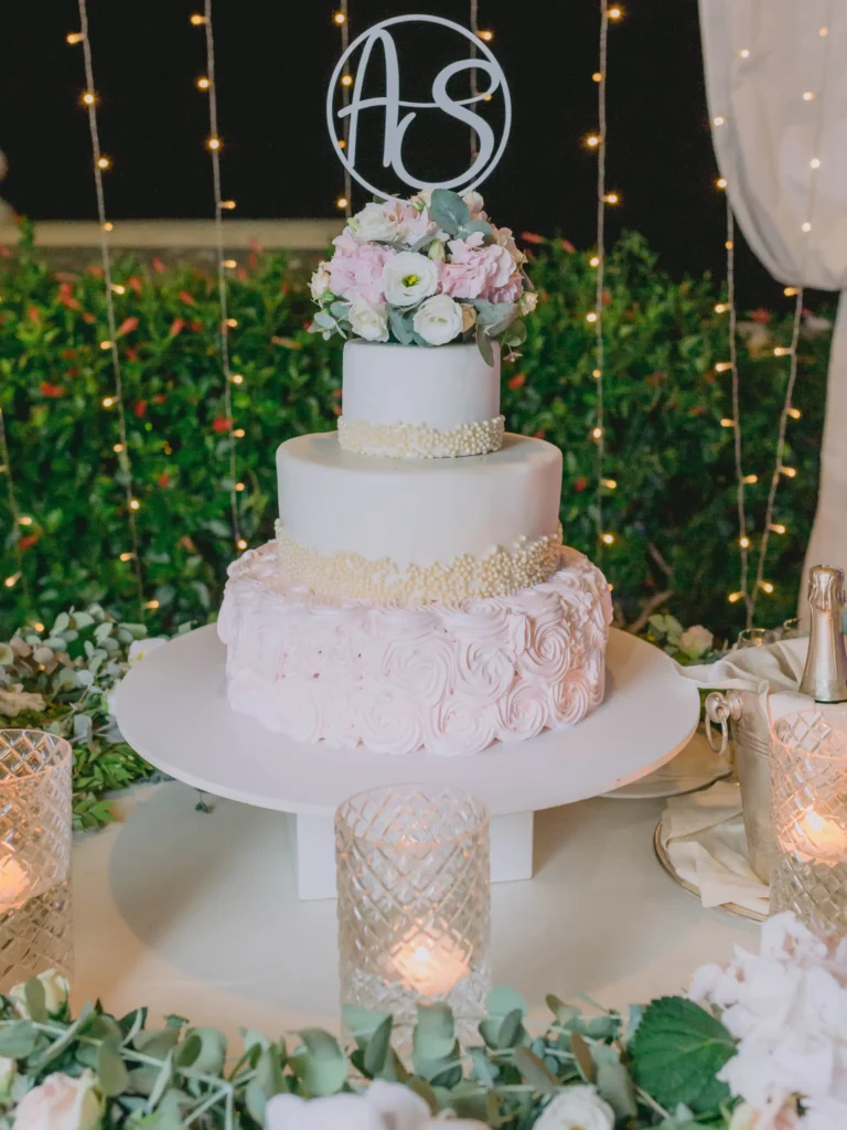 Torta nuziale a tre piani decorata con fiori freschi e un topper personalizzato, circondata da luci e candele romantiche."
