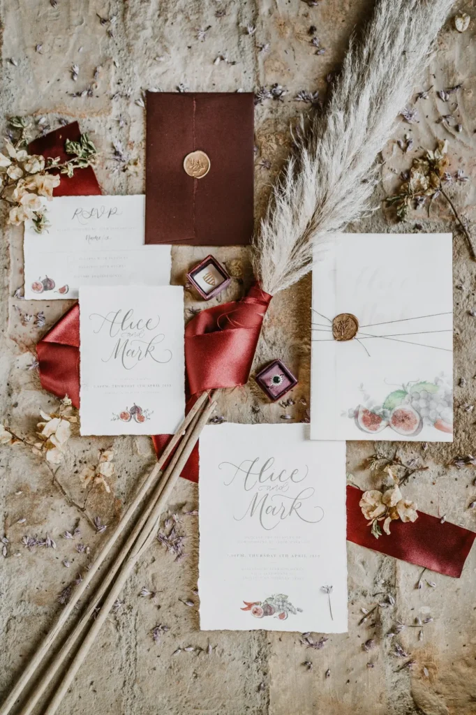 Inviti di nozze decorati in stile rustico con ceralacca, dettagli floreali e fiocchi rossi, adagiati su una superficie naturale.