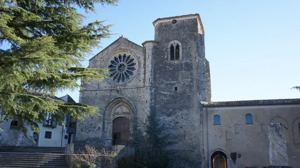 The historic facade of the Church of Santa Maria della Consolazione in Altomonte, a jewel of medieval architecture in Calabria, featuring a large central rose window and a charming bell tower. The setting is framed by trees, creating a picturesque atmosphere ideal for weddings.