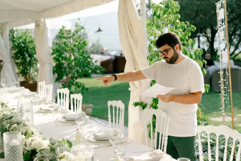 Loris Gallo, in a white T-shirt, points to a detail on a sheet of paper as she inspects the table set for a wedding. The setting is elegant and surrounded by greenery, with white floral decorations that add a touch of sophistication to the event.