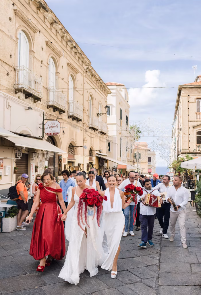 Corteo nuziale con spose e musicisti in un borgo storico